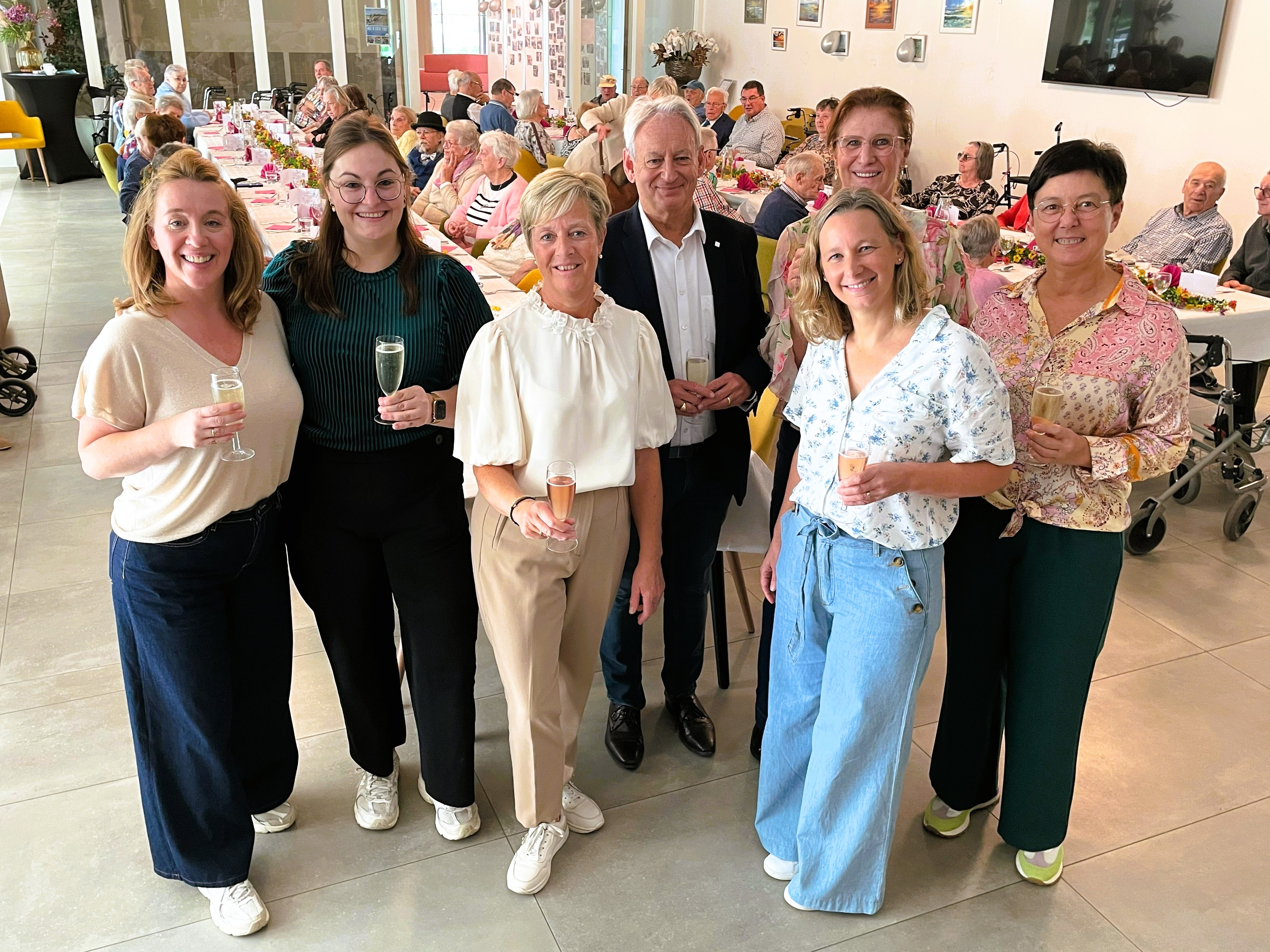 Het team van centrum voor dagverzorging Den Erker heft, samen met algemeen directeur Christian Fillet, het glas naar aanleiding van 25 jaar CDV Den Erker. Op de achtergrond genieten de genodigenden van een gezellige feestmaaltijd.
