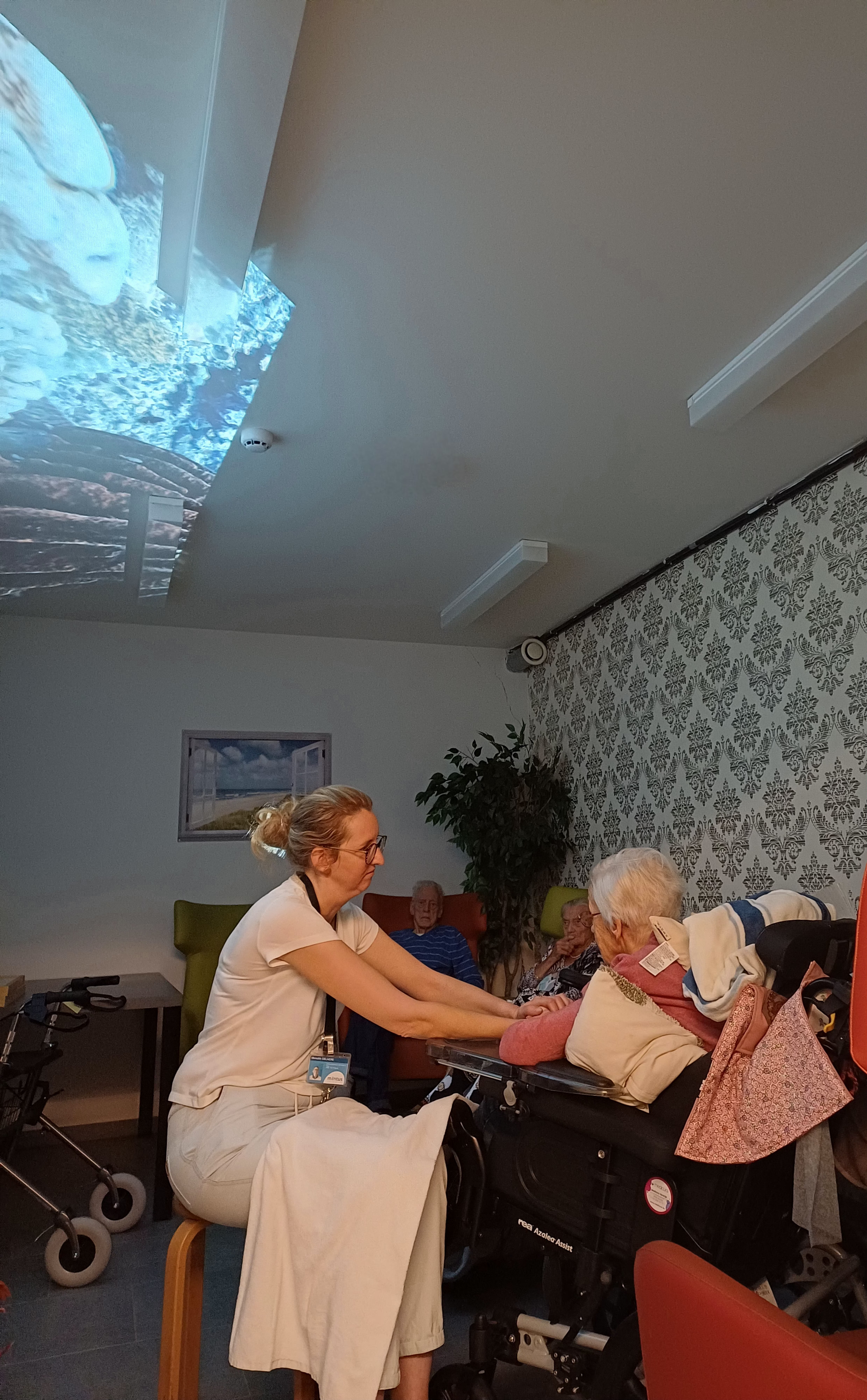 Bewoonster en verzorger zitten samen in de welzijnsruimte met een projectie op het plafond